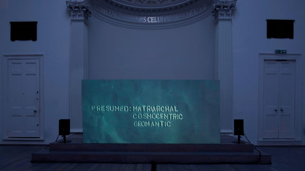 A screen in an old church hall reads "Presumed: Matriarchal, Cosmocentric, Geomantic."