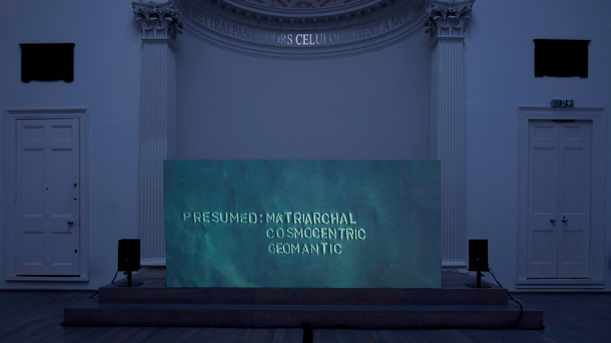 A screen in an old church hall reads "Presumed: Matriarchal, Cosmocentric, Geomantic."